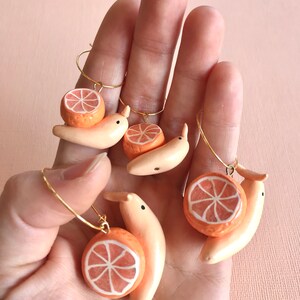Pu&ograve; includere: Quattro paia di orecchini fatti a mano con una fetta di arancia in ceramica e una banana. Gli orecchini sono dorati e hanno un design ad anello.
