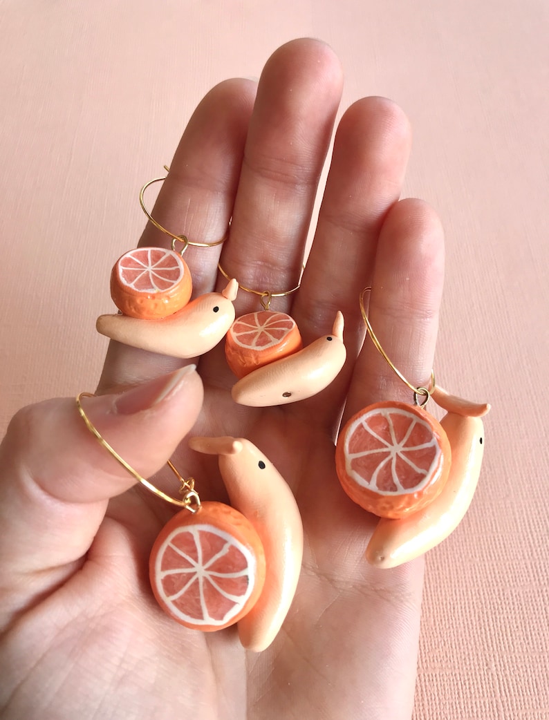 Pu&ograve; includere: Quattro paia di orecchini fatti a mano con una fetta di arancia in ceramica e una banana. Gli orecchini sono dorati e hanno un design ad anello.