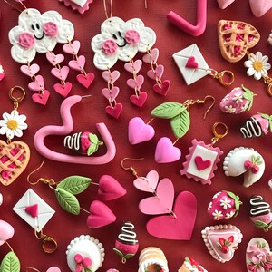 May include: Assortment of pink and white polymer clay earrings featuring various designs, including hearts, clouds, strawberries, cakes, and pies. The earrings are displayed on a red background.