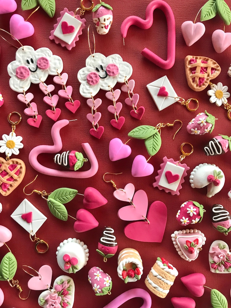 May include: Assortment of pink and white polymer clay earrings featuring various designs, including hearts, clouds, strawberries, cakes, and pies. The earrings are displayed on a red background.