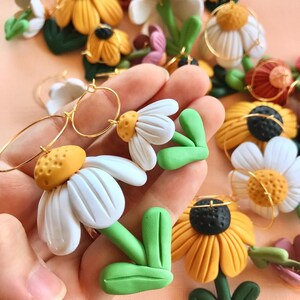 May include: A pair of handmade polymer clay earrings featuring white daisy flowers with yellow centers and green leaves. The earrings are attached to gold hoops.