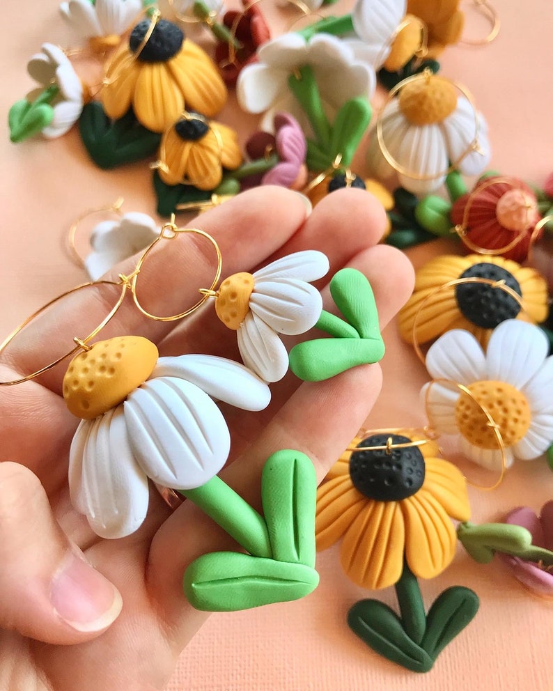 May include: A pair of handmade polymer clay earrings featuring white daisy flowers with yellow centers and green leaves. The earrings are attached to gold hoops.