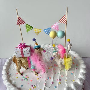 K&ouml;nnte beinhalten: Ein wei&szlig;er Kuchen mit Zuckerguss, verziert mit Lama-Figuren, die Partyh&uuml;te und Federboas tragen. Der Kuchen ist mit einer Geschenkbox, bunten Konfetti und einem Banner mit gemusterten Flaggen und Luftballons geschm&uuml;ckt. Ein festlicher Geburtstagskuchen.