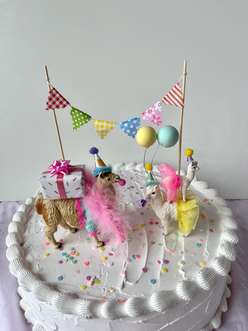K&ouml;nnte beinhalten: Ein wei&szlig;er Kuchen mit Zuckerguss, verziert mit Lama-Figuren, die Partyh&uuml;te und Federboas tragen. Der Kuchen ist mit einer Geschenkbox, bunten Konfetti und einem Banner mit gemusterten Flaggen und Luftballons geschm&uuml;ckt. Ein festlicher Geburtstagskuchen.