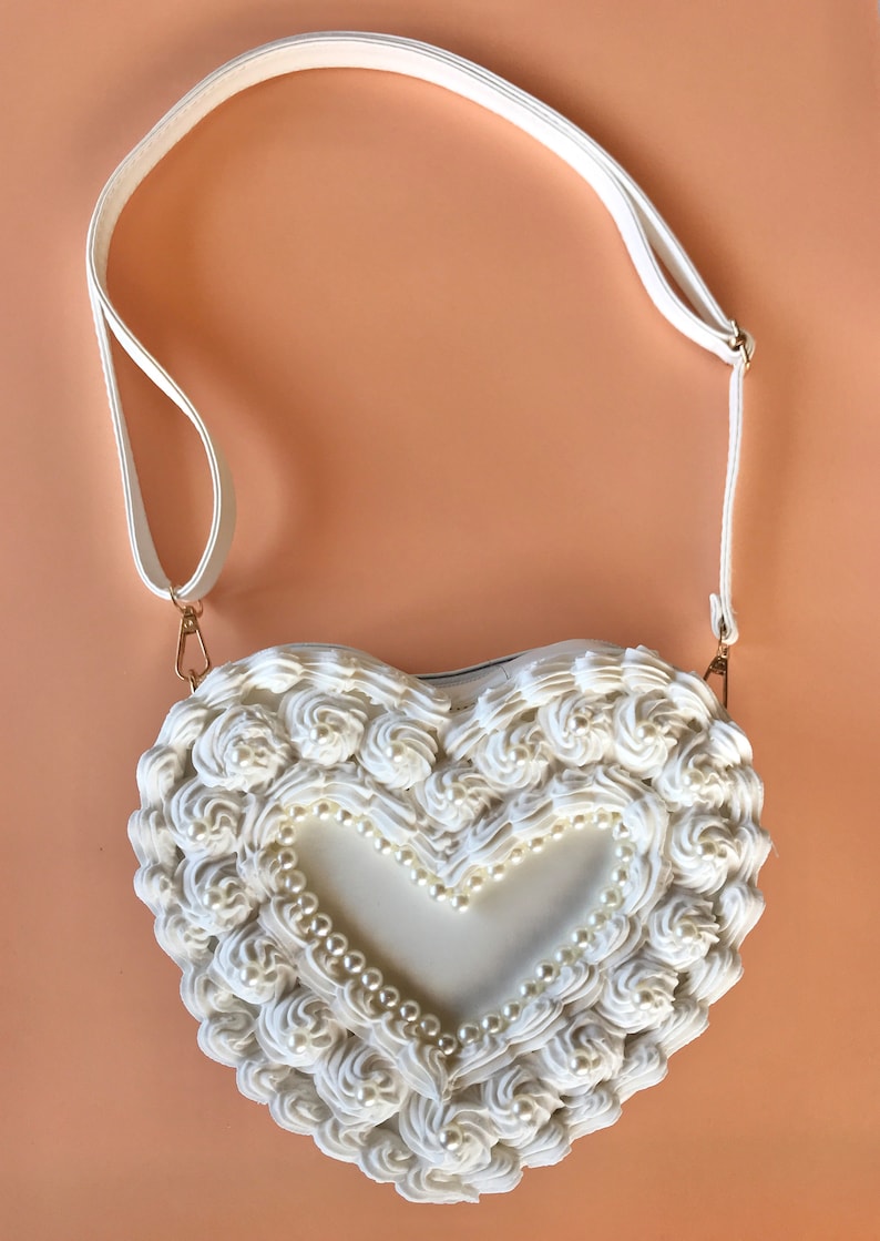 May include: White heart-shaped handbag with a white strap and pearl accents. The bag is made to look like a cake with frosting.