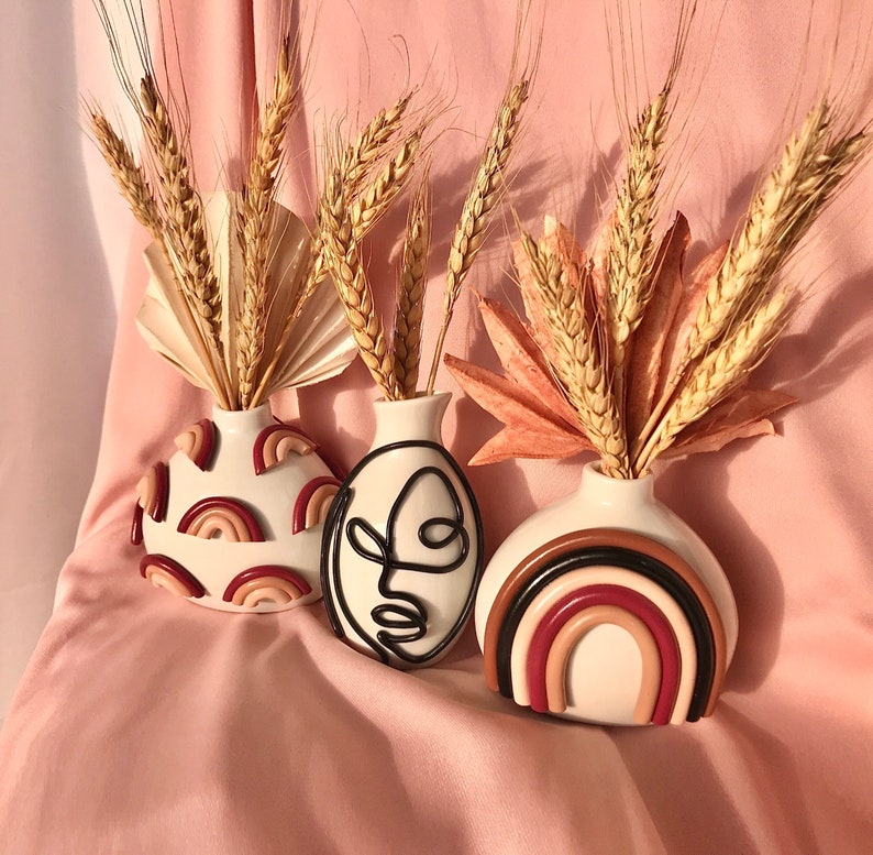 Pu&ograve; includere: Tre vasi in ceramica bianca con diversi design. Il vaso a sinistra ha un design a spirale rosa e rosso. Il vaso al centro ha un disegno a linea nera di un viso. Il vaso a destra ha un design arcobaleno rosa e rosso.