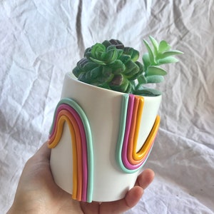 May include: White ceramic planter with a rainbow design. The planter is decorated with three rainbow stripes in yellow, orange, pink, and light blue. The planter is filled with a green succulent plant.