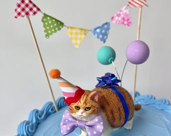 May include: A cake topper featuring a ginger and white cat wearing a party hat and a purple bow tie. The cat is on a blue frosted cake, decorated with a colourful banner and balloons. The cake is a festive decoration.