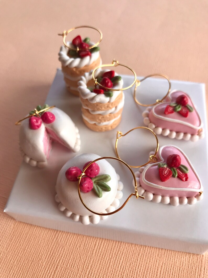 May include: Four pairs of dangle earrings featuring miniature clay cakes and hearts decorated with white frosting, pink icing, and red strawberries. The earrings are made with gold hoops.