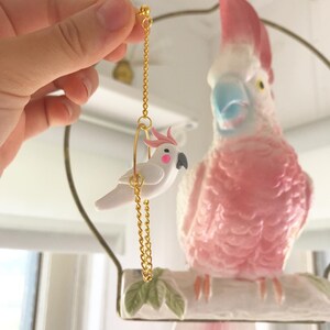 May include: A pair of gold earrings featuring a small white cockatoo on a swing. The cockatoo has pink accents on its crest and beak. The earrings are hanging from a gold chain.
