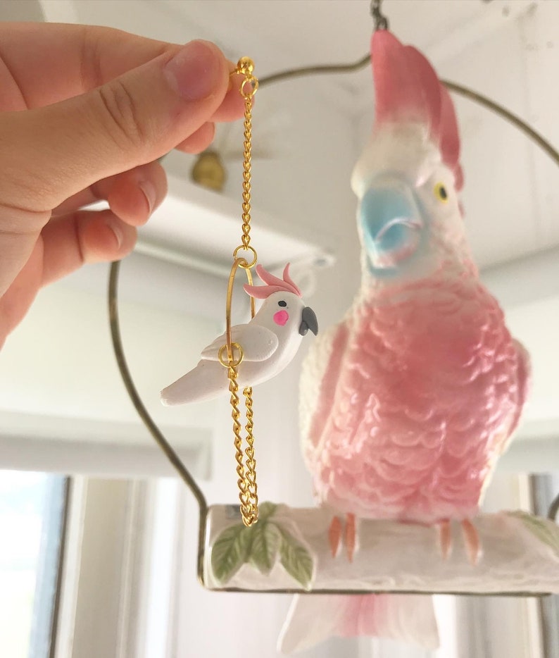 May include: A pair of gold earrings featuring a small white cockatoo on a swing. The cockatoo has pink accents on its crest and beak. The earrings are hanging from a gold chain.