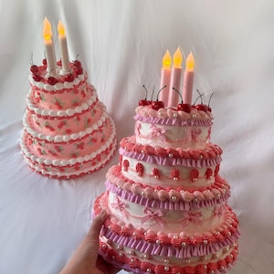 May include: Two tiered cakes decorated with pink and white frosting, cherries, and pearl accents. Both cakes have lit candles on top.