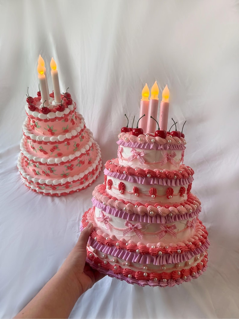 May include: Two tiered cakes decorated with pink and white frosting, cherries, and pearl accents. Both cakes have lit candles on top.