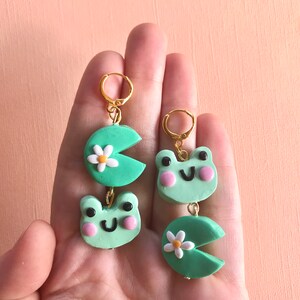 Pu&ograve; includere: Un paio di orecchini pendenti a tema rana verde. Ogni orecchino presenta un viso di rana verde con guance rosa e una ninfea verde con un fiore bianco.