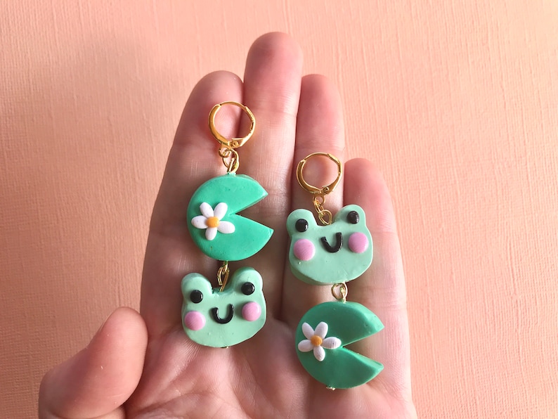 Pu&ograve; includere: Un paio di orecchini pendenti a tema rana verde. Ogni orecchino presenta un viso di rana verde con guance rosa e una ninfea verde con un fiore bianco.