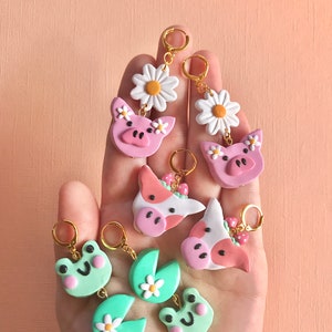 Pu&ograve; includere: Un set di orecchini fatti a mano in pasta polimerica con vari design a tema animali da fattoria e natura. Gli orecchini sono rosa, verdi e bianchi con cerchietti dorati. I design includono un maiale, una mucca, una rana e una margherita.