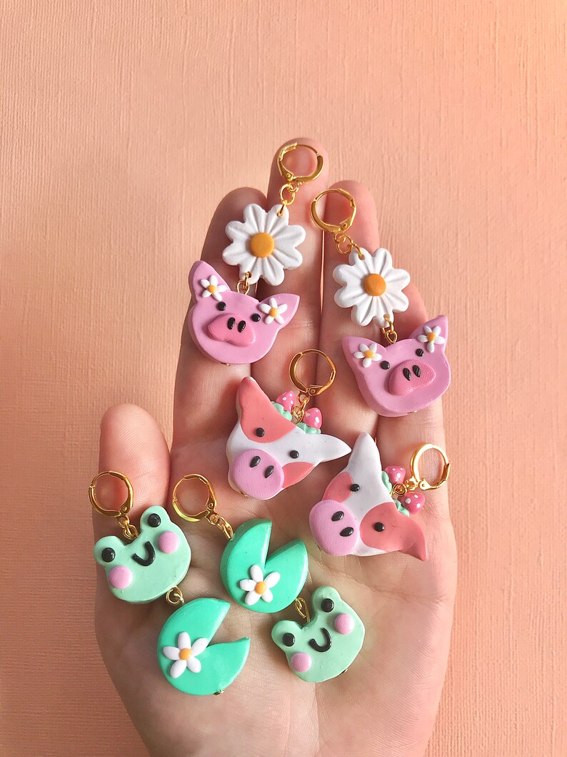 Pu&ograve; includere: Un set di orecchini fatti a mano in pasta polimerica con vari design a tema animali da fattoria e natura. Gli orecchini sono rosa, verdi e bianchi con cerchietti dorati. I design includono un maiale, una mucca, una rana e una margherita.