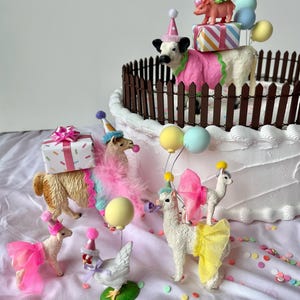 K&ouml;nnte beinhalten: Eine skurrile Geburtstagskuchenszene mit Tierfiguren. Eine Kuh, ein Schwein und mehrere Lamas sind mit Partyh&uuml;ten und bunten Outfits geschm&uuml;ckt. Der Kuchen ist mit einem braunen Zaun verziert und mit Geschenken und Luftballons belegt.