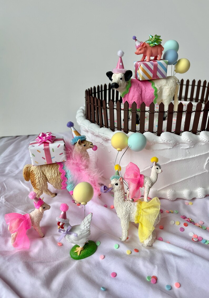 K&ouml;nnte beinhalten: Eine skurrile Geburtstagskuchenszene mit Tierfiguren. Eine Kuh, ein Schwein und mehrere Lamas sind mit Partyh&uuml;ten und bunten Outfits geschm&uuml;ckt. Der Kuchen ist mit einem braunen Zaun verziert und mit Geschenken und Luftballons belegt.