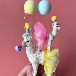 K&ouml;nnte beinhalten: Eine skurrile Figur mit zwei wei&szlig;en Lamas, die mit Partyh&uuml;ten, Tutus und einer rosa Schleife geschm&uuml;ckt sind. Ein Lama h&auml;lt eine Partyhupe, und beide stehen unter pastellfarbenen Luftballons. Eine lustige, festliche Dekoration.