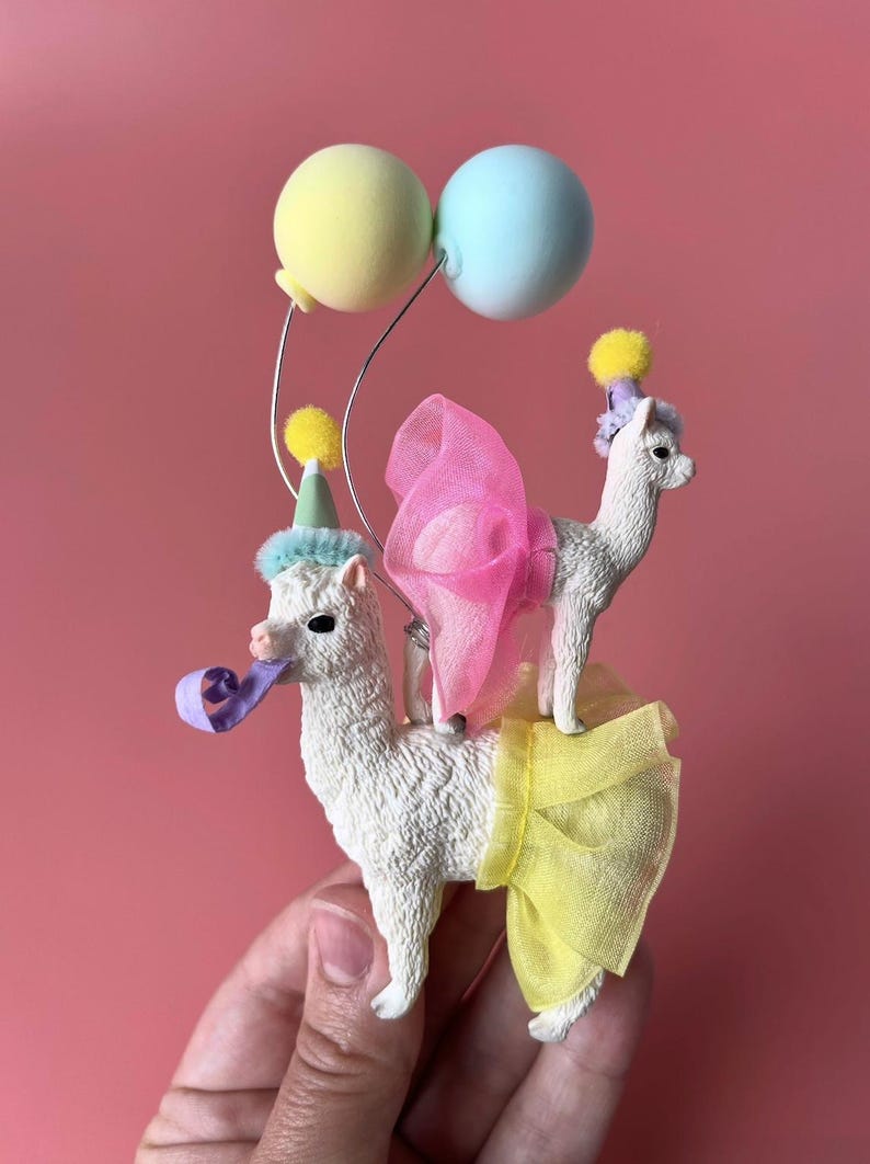 K&ouml;nnte beinhalten: Eine skurrile Figur mit zwei wei&szlig;en Lamas, die mit Partyh&uuml;ten, Tutus und einer rosa Schleife geschm&uuml;ckt sind. Ein Lama h&auml;lt eine Partyhupe, und beide stehen unter pastellfarbenen Luftballons. Eine lustige, festliche Dekoration.