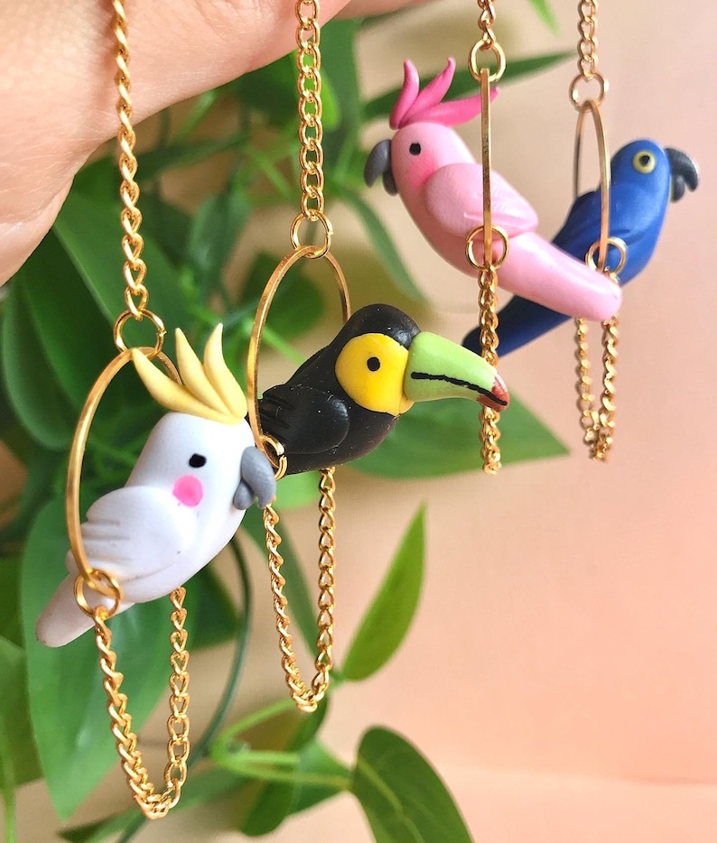 May include: Three colorful bird charms on gold chains. The birds are a pink cockatoo, a black toucan, and a blue parrot. Each bird is attached to a gold hoop.