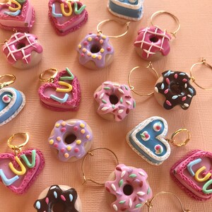 May include: A collection of 16 colorful donut and heart-shaped charms with gold hoops. The donuts are decorated with sprinkles, chocolate, and pink and purple frosting. The hearts are pink and blue with the word "LUV" written on them.