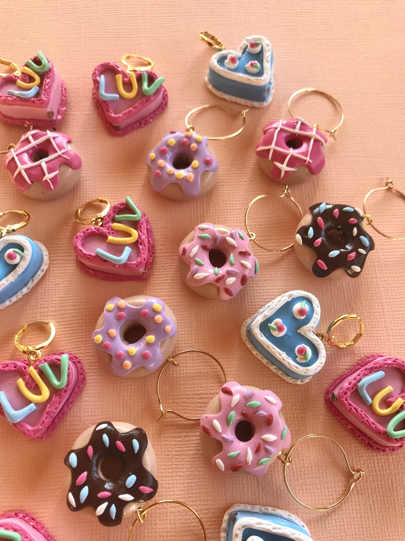 May include: A collection of 16 colorful donut and heart-shaped charms with gold hoops. The donuts are decorated with sprinkles, chocolate, and pink and purple frosting. The hearts are pink and blue with the word "LUV" written on them.