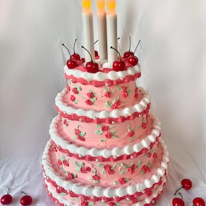 May include: A three-tiered pink cake decorated with white frosting, red cherries, and pearl accents. Three lit candles are on top of the cake.