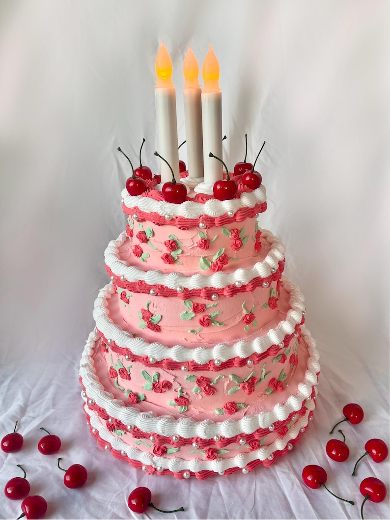 May include: A three-tiered pink cake decorated with white frosting, red cherries, and pearl accents. Three lit candles are on top of the cake.