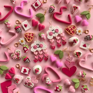 May include: A selection of pink and red polymer clay earrings featuring various shapes, including hearts, clouds, strawberries, and flowers. The earrings are displayed against a pink background.