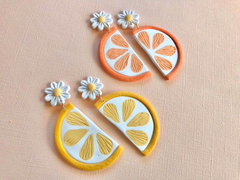 Puede incluir: Dos pares de pendientes hechos a mano. Un par presenta rodajas de naranja con detalles blancos y naranjas, y el otro par rodajas de lim&oacute;n con detalles blancos y amarillos. Cada pendiente tiene un bot&oacute;n de flor de margarita.