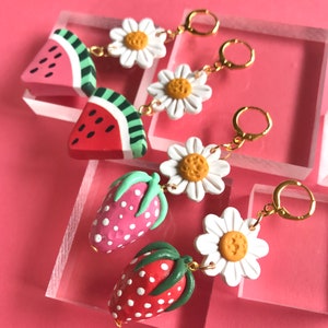 May include: A pair of dangle earrings featuring watermelon and strawberry charms with white daisy accents. The earrings have gold hoops.
