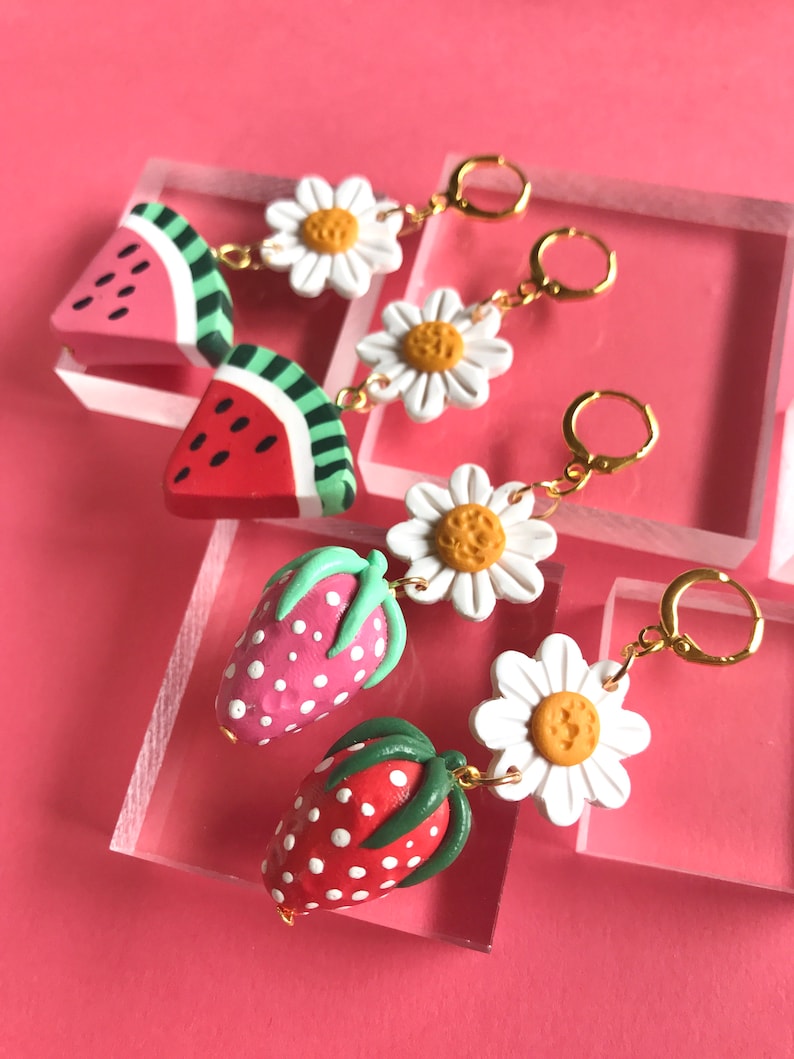 May include: A pair of dangle earrings featuring watermelon and strawberry charms with white daisy accents. The earrings have gold hoops.