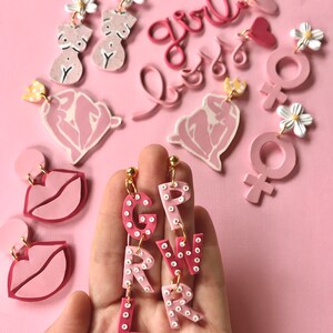 May include: Pink and white polymer clay earrings featuring the word "girl" spelled out in individual letters with white polka dots. Other earrings include a heart, a female symbol, and a stylized female figure.