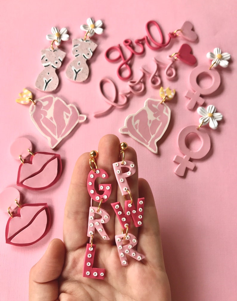 May include: Pink and white polymer clay earrings featuring the word "girl" spelled out in individual letters with white polka dots. Other earrings include a heart, a female symbol, and a stylized female figure.