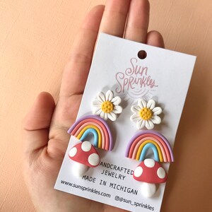 May include: A set of three pairs of handmade polymer clay earrings featuring a rainbow, mushroom, and daisy design. The earrings are made in Michigan and sold by Sun Sprinkles Jewelry. The text on the card reads "Andcrafted Jewelry Made in Michigan www.sunsprinkles.com @sun_sprinkles".