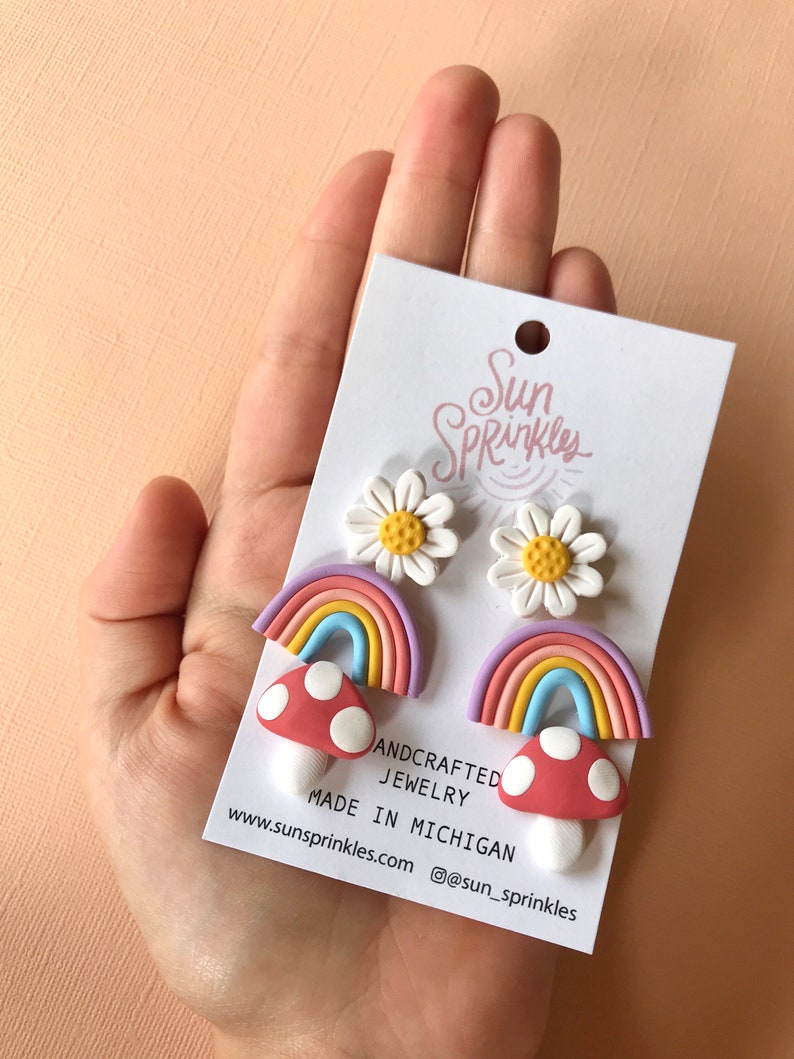 May include: A set of three pairs of handmade polymer clay earrings featuring a rainbow, mushroom, and daisy design. The earrings are made in Michigan and sold by Sun Sprinkles Jewelry. The text on the card reads "Andcrafted Jewelry Made in Michigan www.sunsprinkles.com @sun_sprinkles".
