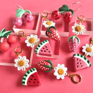 May include: A set of six pairs of dangle earrings featuring various fruit and flower designs. The earrings are made of polymer clay and have gold-toned hooks. The designs include cherries, strawberries, watermelons, and daisies.
