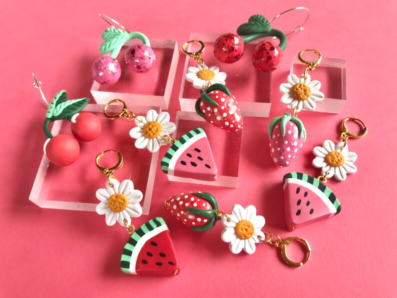 May include: A set of six pairs of dangle earrings featuring various fruit and flower designs. The earrings are made of polymer clay and have gold-toned hooks. The designs include cherries, strawberries, watermelons, and daisies.