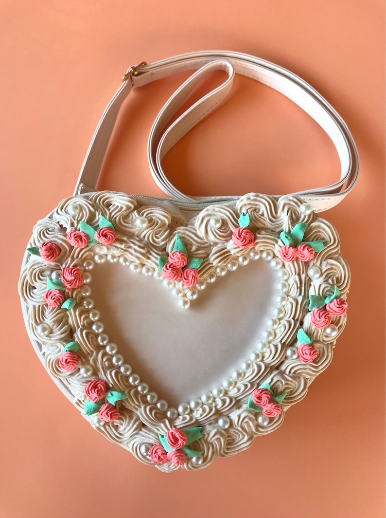 May include: A white heart-shaped handbag with a cream-colored, pearl-embellished, and floral design. The bag has a white strap and a zipper closure.