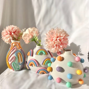 May include: Three ceramic vases with pastel rainbow designs. The vases are decorated with raised, colorful shapes and are filled with pink flowers.