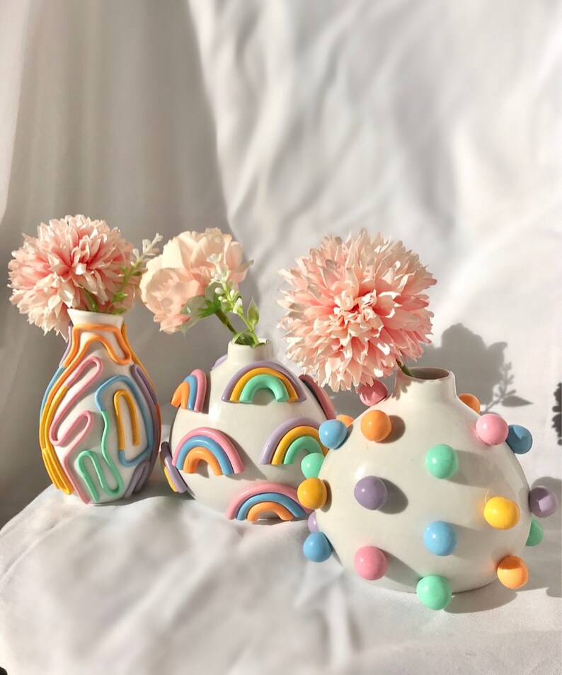 May include: Three ceramic vases with pastel rainbow designs. The vases are decorated with raised, colorful shapes and are filled with pink flowers.