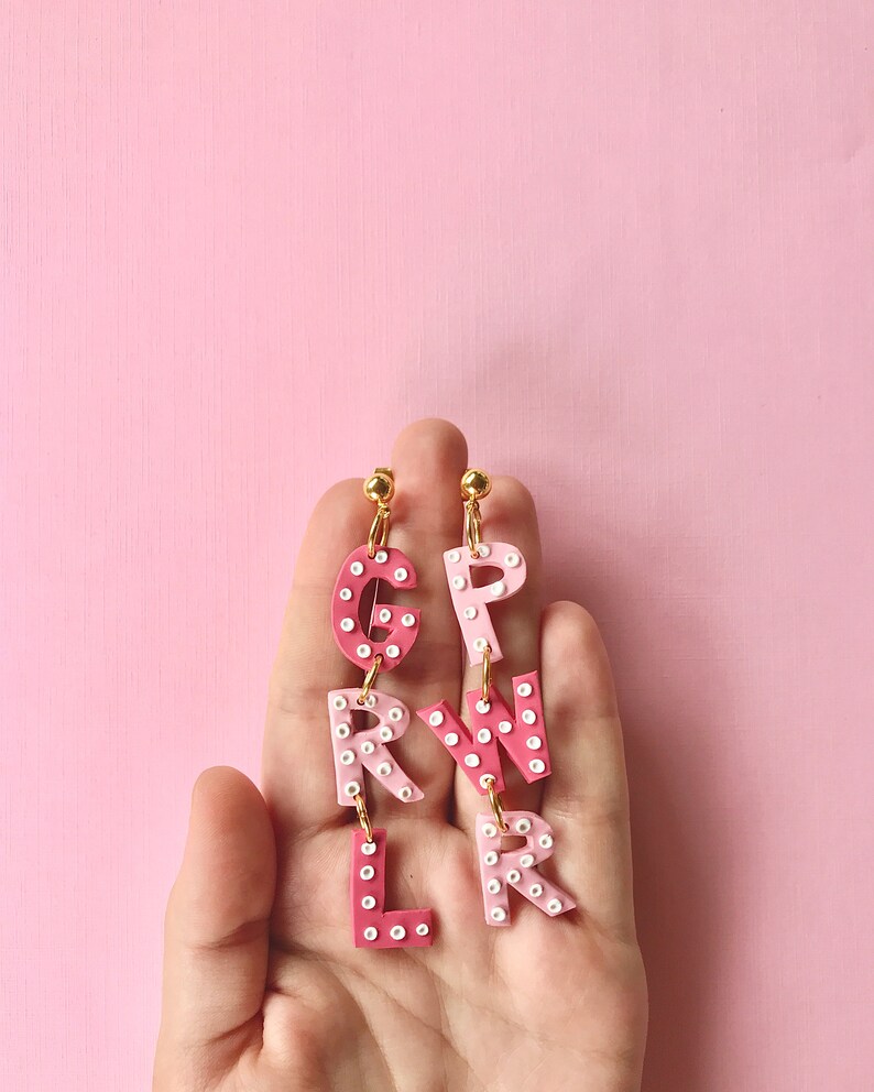 May include: Pink and white letter earrings spelling out "GIRL" with white polka dots. The earrings are hanging from a person's hand.