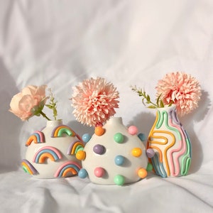 May include: Three ceramic vases with pastel rainbow designs. The vases are decorated with colorful dots, swirls, and rainbows. The vases are filled with pink flowers.