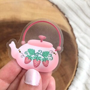 May include: A clear sticker with a pink teapot with a strawberry and leaf design. There are two bees on the sticker.
