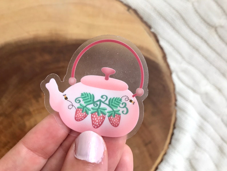 May include: A clear sticker with a pink teapot with a strawberry and leaf design. There are two bees on the sticker.