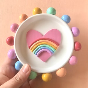 Puede incluir: Un plato de cer&aacute;mica blanca para baratijas con un coraz&oacute;n rosa que contiene un arco&iacute;ris. El plato est&aacute; rodeado por un anillo de bolas de colores pastel.