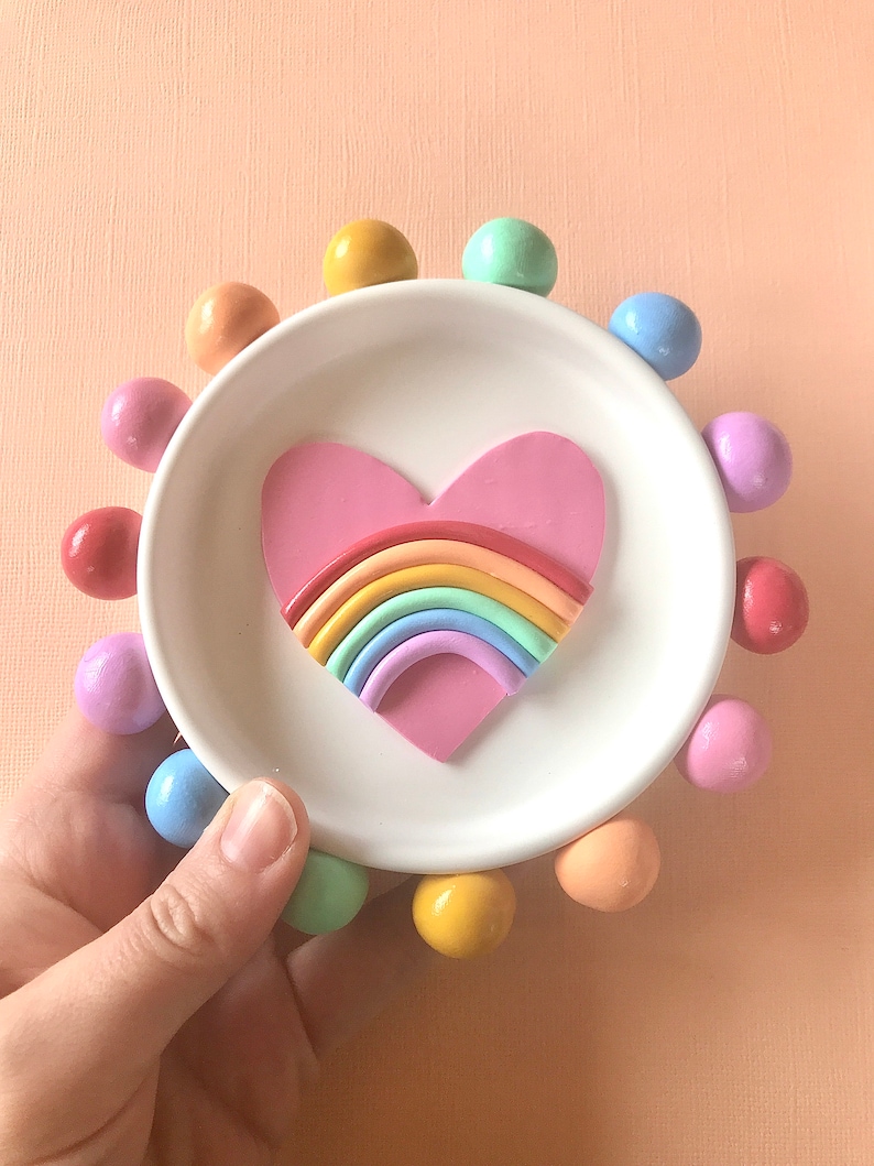 Puede incluir: Un plato de cer&aacute;mica blanca para baratijas con un coraz&oacute;n rosa que contiene un arco&iacute;ris. El plato est&aacute; rodeado por un anillo de bolas de colores pastel.