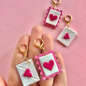 May include: A pair of earrings featuring a pink and white envelope with a pink heart design. The earrings are made of polymer clay and have gold hoops.